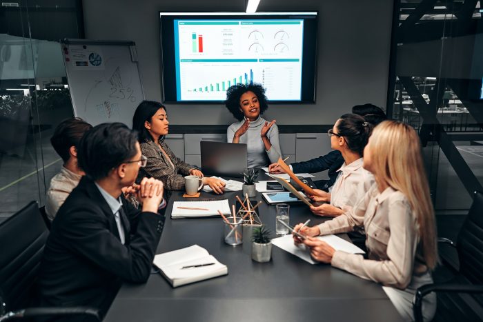 Group of professionals in a boardroom watching a presentation on a wall-mounted screen with charts behind them in a meeting room.