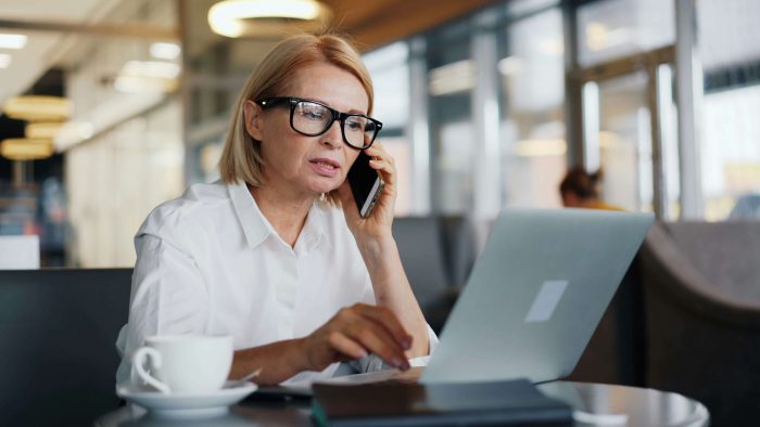 Blonde woman with black glasses in a white shirt sits at a desk, talking on a smartphone in a bright office.