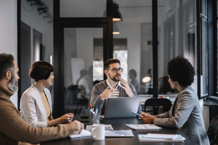 Team meeting around a conference table as a man presents with a laptop beside him in a modern office environment.